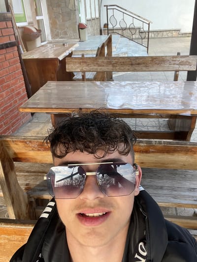 The image depicts a close-up view of a young male situated in an outdoor seating area in Krivodol, Bulgaria. The subject has dark, curly hair and is wearing large, hexagonal-framed sunglasses with a gold-colored bridge. He is dressed in a black padded jacket, with white text visible on the collar. His mouth is slightly ajar, revealing upper teeth, and he has a visible, light mustache.

In the background, behind the subject, is an outdoor patio or terrace furnished with multiple rustic wooden tables and benches. The table immediately in front of the subject is covered with a clear plastic sheet. To the left, a section of a red brick wall is visible. Further back, a light-colored building with stone cladding and white walls is present, featuring a window with green trim and a brown cylindrical waste bin nearby. A decorative metal railing with curved patterns flanks a set of steps leading upwards on the right side of the background. Reflections in the subject's sunglasses show bare tree branches against a bright, partly cloudy sky, indicating an outdoor setting.