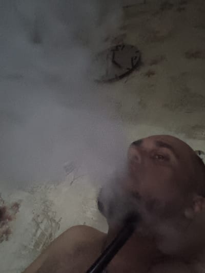The image depicts a close-up, low-angle view of a person exhaling a cloud of smoke, obscuring much of the scene. The subject appears to be a bald individual with dark eyebrows, and a portion of their face and chest is visible. They are holding a dark object to their mouth, from which the smoke is emanating. The background shows a textured, dimly lit ceiling with what appears to be an old, circular fixture or marking on it. The wall beneath the ceiling has a mottled, aged appearance with some reddish-brown stains and what may be faded markings or repairs. The lighting is subdued, creating a hazy and somewhat claustrophobic atmosphere. There is no discernible information within the image to associate it with the city of Zuevka, Russia, or any specific location.
