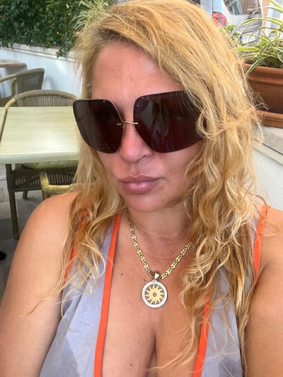 A close-up, over-the-shoulder selfie taken outdoors shows a blonde woman with curly hair wearing large dark sunglasses, an orange and gray tank top, and a gold chain with a sun pendant. She is seated at an outdoor cafe with tables and chairs visible in the background. The environment suggests a warm climate, possibly a seaside town. The location is Đenovići, Montenegro.