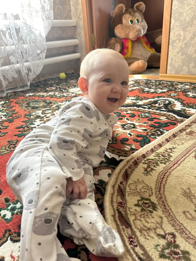 A baby wearing a white onesie with a koala pattern is sitting on a carpeted floor. The baby is smiling and looking towards the camera. In the background, a plush toy bear is sitting on a shelf. The room appears to be a child's bedroom. The carpet has a red and green patterned design.