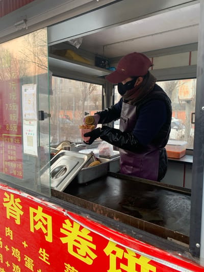 A person wearing a hat, apron, and gloves works at a food stand. The stand features a sign with red and yellow Chinese characters and a price list offering items like "Spicy Chicken Willow" for 7 yuan and "Fried Sausage" for 3 yuan. QR codes are visible on the stand, along with prices for various fillings such as red bean paste at 2 yuan. The worker appears to be preparing food, possibly wrapping it. A metal grill and other cooking equipment are nearby.