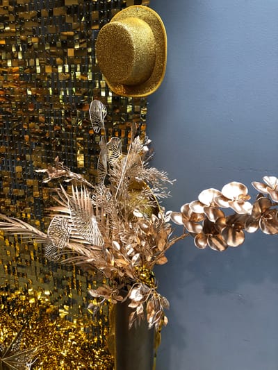 This is a still life image of a festive decoration against a textured blue wall. The left side of the image features a shimmering gold sequin backdrop with a gold glittered top hat hanging slightly off-center and tilted. Below the hat, a bouquet of gold metallic artificial foliage is arranged in a dark vase. A branch with gold metallic orchid-like flowers extends from the bouquet towards the right side of the image. The bottom left corner shows a scattering of gold metallic fringe and a metallic star ornament. The overall aesthetic suggests a celebratory or holiday theme. There are no people or animals present in the image. The location in Vaughan, Canada cannot be determined from the image itself.