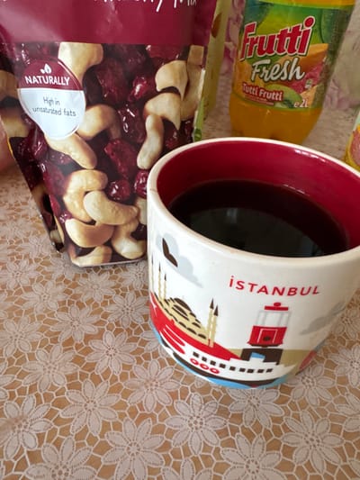 A close-up, elevated view captures a collection of food and beverage items arranged on a table covered with a white lace-patterned tablecloth over a light brown surface, located indoors in Ciorescu, Moldova. A clear plastic bag of "NATURALLY High in unsaturated fats" cranberry mix with visible cashews and dried cranberries stands prominently on the left. To its right, a white ceramic mug with a red interior contains a dark liquid. The mug's exterior features a multi-colored graphic illustration depicting a red tram, a ferry boat, and a mosque-like structure, with the red text "İSTANBUL" above the tram. Behind these items, a transparent plastic bottle of "Frutti Fresh Tutti Frutti" beverage, labeled as 2L and containing an orange liquid, is visible. No individuals are present in the scene.