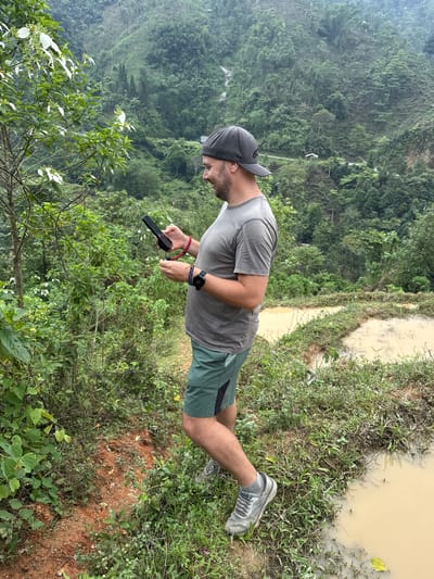 The image depicts a man standing on a dirt path next to a water-filled agricultural terrace, set against a backdrop of lush green mountains. The scene is characteristic of a mountainous, vegetated region, specifically in Nậm Dịch Commune, Vietnam.
The man, positioned on the right side of the frame, is seen in profile, looking down. He wears a grey baseball cap, a plain grey t-shirt, green and black shorts, and grey sneakers. Accessories include a black smartwatch on his left wrist and red and blue bracelets on his right wrist. He holds a black rectangular electronic device, possibly a smartphone or small camera, which is connected by a red cord. His posture suggests he is focused on the device in his hands.
To the man's right, a muddy, shallow body of water, likely part of a terraced rice paddy system, extends along the edge of the path. The banks of this water feature are overgrown with various green plants and grasses, and small dark specks, possibly insects, are visible on the water's surface. In the background, dense, verdant mountains covered in trees and foliage rise, appearing somewhat hazy in the distance. The foreground also features various green plants and bushes.
The time of day appears to be daytime, possibly overcast or with soft light, as strong shadows are absent. The overall impression is one of a remote, natural environment with agricultural elements. There is no visible text in the image.
