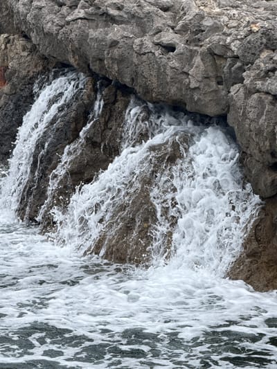 This image shows a natural waterfall cascading down rough, grey rocks into the sea. The water is frothy and white as it tumbles and splashes against the rocks and into the dark, choppy water below. The rocks are textured and uneven, with crevices and overhangs. The scene appears to be located along a rocky coastline. This is a view of the rocky coast in Santander, Spain, specifically likely near the Sardinero area where the urban landscape meets the sea. There are no people or man-made objects visible in the immediate scene. The focus is on the power and motion of the water and the natural ruggedness of the rocks.