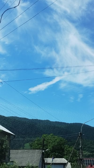 The image shows a view of a mountainous landscape with a bright blue sky filled with wispy white clouds. Several dark electrical wires crisscross the frame horizontally. In the foreground, the roofs of two buildings are visible, one with a corrugated grey roof and the other with a lighter-colored roof. The buildings appear to be situated at the base of a densely forested mountain. A utility pole with a street lamp is also present on the right side of the image. The scene is located in Mizhhiria, Ukraine, as indicated by the context provided.