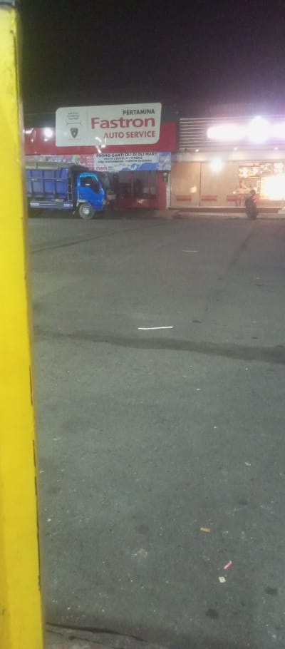 A Pertamina Fastron Auto Service station, advertising "Promo Ganti Oli di Oli Mart" (Oil Change Promo at Oli Mart). A blue truck sits outside, possibly for service. A Dunkin' Donuts shop appears adjacent to the auto service. The scene is dimly lit, likely at night. Litter is visible on the ground. A yellow vertical element, possibly a pole, is present on the left side of the frame.