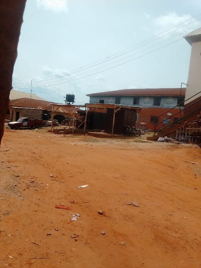 The image shows a dirt road in front of a building with the sign "Startimes" on it, located in Auchi, Nigeria, at coordinates 7.05 latitude and 6.263 longitude. The building has a red roof and is in the background of the image. There is a small wooden structure in front of the building. There is a pickup truck parked on the dirt road. The road is covered in dirt and debris. The sky is clear and there are no people in the image. The building is in good condition, and the wooden structure appears to be in working order.