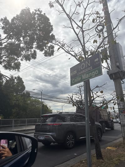 A street sign in San Rafael, Costa Rica (latitude: 9.937, longitude: -84.152) reads "ION CHARGERS Calle Mango 1.7 km."  The sign is mounted on a pole with a utility box. A dark gray SUV with the word "PATHFINDER" on the back is stopped in traffic.  The vehicle is facing right.  The SUV is in the right lane of the road. A dump truck is in the left lane. Both vehicles are facing right.  There is a thin tree in the foreground on the right.  Behind the tree, there is a streetlight.  The tree in the background is bare.  The sky is overcast.  The road is paved with black asphalt.  There is a metal fence in the background, to the left of the road.  A hand is holding a phone, partially visible in the lower left of the frame.  There is a car in the background.  A person is holding a phone in the lower left corner of the frame.
