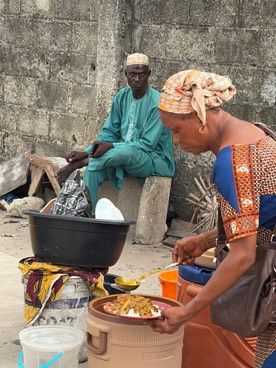 An outdoor scene in Jakande, Nigeria, features two individuals and an informal food vending operation. In the foreground, a woman with dark skin and a patterned beige headwrap is actively serving food. She wears a blue top with patterned orange and brown sleeves and a brown shoulder bag. Her right hand holds a golden ladle, scooping from a container, while her left hand supports a red plate containing white rice topped with a brown stew with meat pieces. The plate rests on a beige "Pot Trendy" insulated food container, adjacent to a blue container. To her left, a large black plastic basin filled with miscellaneous items, including a black plastic bag, sits atop two stacked white/grey buckets draped with a yellow and red patterned fabric. A clear plastic bucket is visible on the ground.
In the background, a man with dark skin and a teal-colored traditional garment (tunic and trousers) is seated barefoot on a concrete block. He wears a light-colored embroidered cap and has a visible mark on his forehead. His hands are clasped in his lap, possibly holding a small dark object. He looks directly forward with a serious expression. Behind him is a textured, weathered grey concrete block wall. The ground is uneven, composed of dirt and debris, with a wooden stool and some scattered sticks visible near the man.