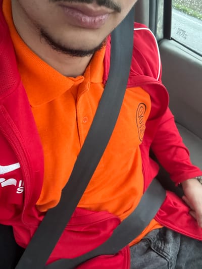 A partial view of an individual's upper torso and lower face is presented, seated in a vehicle. The scene is located in Illkirch-Graffenstaden, France. The individual exhibits a mustache and a dark chin-strap beard. The lips are slightly parted. They are attired in an orange polo shirt featuring a dark, stylized ear-like logo with partial text on the right chest area, layered under a red zippered jacket with white trim and partial letters "IS" on the left chest. A black vehicle seatbelt traverses diagonally across the torso and lap. The interior of the vehicle is visible, with a passenger-side window displaying a blurred exterior scene suggestive of wet weather conditions. A wrist, adorned with a bracelet or watch, is visible on the right side of the frame. The lower body shows dark-colored pants, possibly denim.