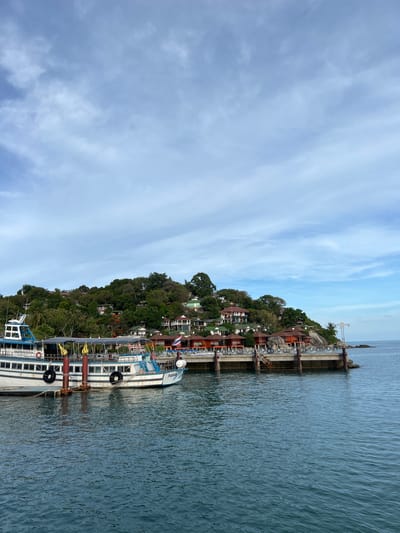 In Ban Haad Rin, Thailand, a docked ferry with the name "HAADRIN BUS BUDDHA" visible on its side is moored at a pier. The ferry, primarily white with blue accents, has a covered seating area with a grey canopy and two decorative golden flame-like structures. Several black tires serve as fenders along its side.
The pier, constructed of concrete and metal, extends into the calm, blue water. Along the pier, a row of buildings with orange and red roofs is visible, nestled into a lush, green hillside. These buildings appear to be part of a resort or guesthouse, with balconies and walkways overlooking the sea. A Thai flag flies from one of the structures.
The hillside is densely covered with trees and foliage, with some houses interspersed among them, suggesting a coastal community built on an incline. The sky above is a mix of light blue with wispy white clouds. The overall scene depicts a tranquil coastal scene in a popular tourist destination.