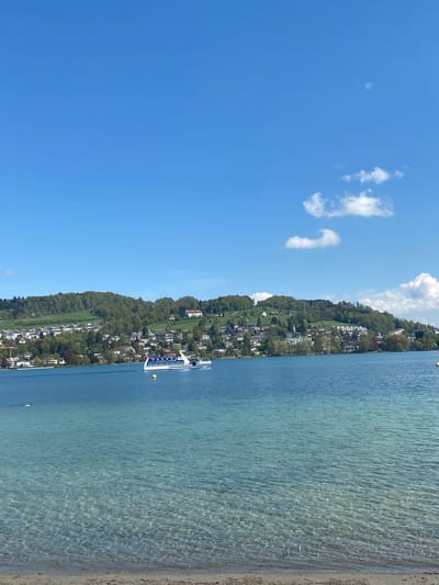 This media file captures a picturesque and serene landscape, likely Lake Lucerne in Switzerland, under a clear blue sky. The image is composed of three main horizontal bands: a vast expanse of bright blue sky at the top, a body of clear water in the middle, and a lush green hillside dotted with buildings to the left, which slopes down to meet the water. A narrow strip of sandy or pebbly shoreline is visible on the far right, where the water meets the land.

The sky is a vibrant azure, with a few scattered, wispy white clouds adding texture. The water is a striking light blue-green, reflecting the sunlight and showing intricate ripple patterns, particularly in the shallower areas near the shore. The transparency of the water suggests excellent clarity.

On the left side of the lake, a verdant hillside is densely covered with trees and numerous white-colored buildings, possibly homes or hotels, interspersed among the greenery. This suggests a populated lakeside community nestled into the landscape.

A prominent object in the water is a sleek, white passenger boat with blue accents along its upper deck, which appears to be cruising calmly on the lake. A yellow buoy floats in the water nearby. No discernible text is visible on the boat or elsewhere in the image.

The scene depicts a beautiful, sunny day, likely mid-morning or early afternoon, with calm weather. The overall impression is one of tranquility and natural beauty, characteristic of a scenic European lake setting like Lucerne.