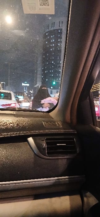 The image depicts a nighttime urban scene in Makkah Al Mukarramah, Saudi Arabia, viewed from inside a vehicle through its front windshield. The windshield appears smudged or wet, and a QR code sticker is affixed to its upper right portion. The vehicle's dark-colored dashboard, an air vent, and a partial view of the right-side A-pillar are visible in the foreground.
In the midground, a woman dressed in a black niqab stands on a street or sidewalk, holding an infant or small child wrapped in a light pink blanket or fabric against her chest. The child's face is partially visible. Multiple vehicles are present on the road, with a white sedan prominent on the left. Green traffic signals and streetlights illuminate the area. In the background, a tall, multi-story building with numerous illuminated windows and a distinctive architectural top is visible, characteristic of an urban high-rise.