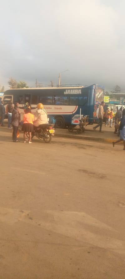 A blue and white bus, labeled "JAMBA" and "Lopha Travels," is parked on a street with people nearby. A motorcycle with two people sits in front of the bus. Other individuals are scattered around, some walking, some standing. A small cart with boxes sits next to the bus. The sky is overcast. The road is an orangey tan color.