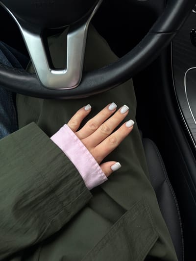 A person's hands with white French manicured nails are visible in the driver's seat of a car. The person is wearing a dark green jacket over a pink long-sleeved shirt. The car's steering wheel and dashboard are also visible. The scene appears to be in Dreiliņi, Latvia, a location known for its shopping centers and residential areas.