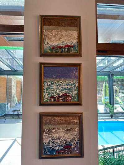 Three framed paintings are displayed vertically on a light-colored wall. The paintings feature abstract depictions of small houses with red roofs. The top painting has a background of horizontal stripes in shades of brown and white, with white paint splattered across it. The middle painting features a dark blue sky above a choppy green and white sea, with small houses at the bottom. The bottom painting is similar to the top one, with a brown and white abstract background and splattered white paint.
To the left of the paintings, large windows reveal an indoor/outdoor space with a glass roof. Outdoor furniture, including blue chairs and a small table, are visible. The floor is tiled.
To the right of the paintings, a bright blue swimming pool is visible through large windows. Lush green plants and outdoor seating are also present. The overall environment suggests a modern residential or resort setting, possibly in Panichishte, Bulgaria, given the context. There are no people visible in the image.