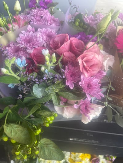 A vertical, close-up, eye-level shot captures a bouquet of vibrant flowers. The bouquet is predominantly filled with pink roses and purple chrysanthemums, interspersed with delicate blue and white blossoms and sprigs of tiny purple flowers. Green foliage and branches with small, green berries add a natural touch to the arrangement. The flowers are partially wrapped in translucent purple and white paper, adding a soft, diffused quality to the image. The background is blurred, with hints of other flowers and a dark, reflective surface. The overall lighting is soft and diffused, creating a dreamy and ethereal effect. The image is taken in Hadera, Israel.