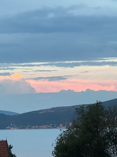 The image captures a panoramic view of Baošići, Montenegro, during what appears to be twilight. The sky is a dominant feature, displaying layers of clouds in various shades of blue and gray, with streaks of soft pink and yellow indicating the lingering light of sunset or sunrise. Below the sky, a range of dark, forested hills slopes down towards a body of calm, light blue water. Along the shoreline of the hills, a scattering of lights and buildings suggests a settlement within the town. In the foreground, the dark silhouette of trees frames the scene, with a portion of a reddish-brown tiled roof visible in the lower left corner, hinting at residential architecture. The overall impression is one of serene natural beauty, with a human presence subtly integrated into the landscape.
