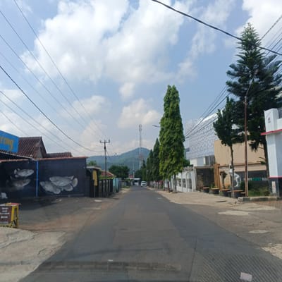 This is a ground-level, eye-level, forward-facing medium shot of a street in Babatan, Indonesia. The street is paved and appears to be a two-lane road with some minor damage and patches. On the left side of the road, there is a dark-colored building with a mural of fish painted on its exterior. A blue sign with yellow text is partially visible above the mural. Adjacent to this building are fences and houses with tiled roofs. Further down the street on the left, there are more fences and buildings.
On the right side of the road, there is a white building with elaborate patterned walls and columns. Several tall, dark green evergreen trees are planted along the sidewalk. A white statue of a horse is also visible. Further down the street on the right, a modern white building with a red trim is present.
The sky is blue with scattered white clouds. Several power lines are strung across the sky, supported by poles. In the distance, a mountain range is visible, with a tall communication tower situated on one of the peaks. A white vehicle is driving down the street in the middle distance. The overall environment suggests a semi-urban or suburban area with a mix of residential and commercial buildings.