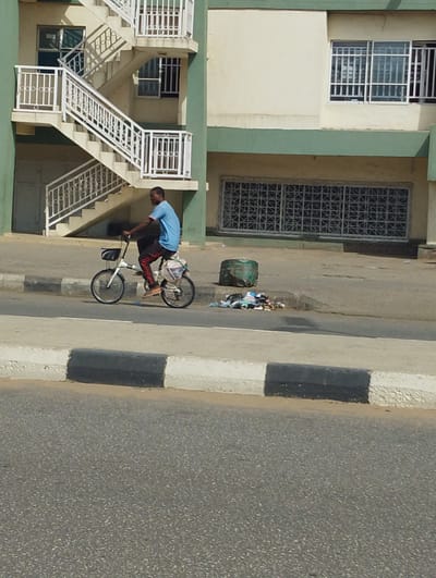 The image captures a scene on a street in Babban Giji, Nigeria. A man is riding a white bicycle along the sidewalk. The man is dark-skinned, and wearing a light blue t-shirt and dark pants. He is carrying something on the back of his bicycle. The sidewalk is edged with a black and white curb. Next to the sidewalk, a wrapped green trash can is sitting, and next to it, garbage is strewn along the curb. Behind the sidewalk is a building. The building has external white stairs with white railings. The building is painted white with green accents. There are barred windows. The street is paved asphalt.