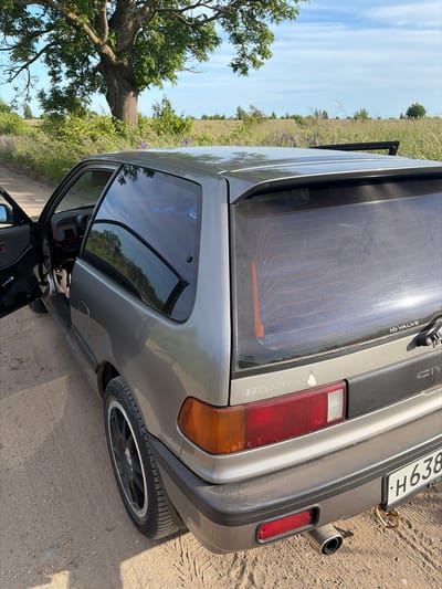 A gray, two-door Honda Civic hatchback is parked on a dirt road, facing right, with its driver's side door open. The car is positioned in a rural landscape with a large tree to the left, green vegetation, and a blue sky with some clouds. The rear of the car is visible, showing a red taillight, a black bumper, a silver exhaust pipe, and a license plate that reads "H638". A small sticker of a white pine tree is visible on the rear of the car. The scene appears to be set in a natural environment, possibly outside the city of Povarovka, Russia, given the context.
