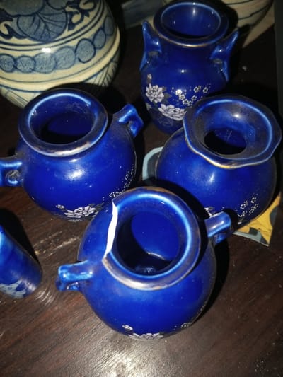 The image shows several small, blue ceramic vessels. They appear to be vases or jugs, some featuring white floral designs. One of the vases has a crack on its rim. They are arranged on a dark wooden surface, with a larger blue and white patterned ceramic object partially visible in the background. The lighting is somewhat dim, adding shadows to the scene.