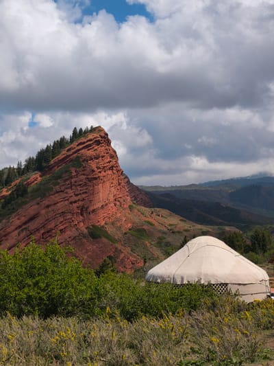 This vertical shot captures a dramatic natural landscape in Jeti-Ögüz, Kyrgyzstan, featuring a striking red rock formation and a traditional yurt. The central subject is an imposing, craggy mountain on the left, composed of distinct horizontal layers of rich red rock. Its upper slopes and jagged crest are adorned with dense evergreen trees, likely pines or firs, providing a contrasting dark green against the reddish hue.
In the foreground on the right, a cream or white canvas yurt is partially visible, nestled amidst lush green bushes and wild shrubs. The yurt's characteristic domed shape and a section of its intricate wooden lattice frame (kerege) can be discerned through the surrounding foliage. The immediate foreground is dominated by these green bushes, some displaying small clusters of yellow flowers, adding a touch of color to the lower portion of the image.
Beyond the prominent red rock, the midground and background reveal rolling hills and distant mountains, covered in a mix of green vegetation and some barren patches. The sky overhead is largely covered by an expansive array of grey and white clouds, suggesting an overcast or changeable weather condition, though patches of bright blue sky are visible in the upper left. The diffused daylight indicates it is daytime, possibly late morning or early afternoon. No people or visible text are present. The scene evokes a sense of rugged natural beauty and traditional nomadic culture in a remote mountainous setting.