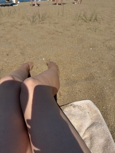The image shows a close-up view of a person's legs and feet on a sandy beach. The person is lying down on a towel, and their legs are extended. In the background, other people can be seen walking on the beach near the water. The environment is a beach with sand and sparse vegetation. The scene appears to be a sunny day. The location is not specified as Антонов ауылдық округі, Kazakhstan, nor can it be inferred from the image itself.