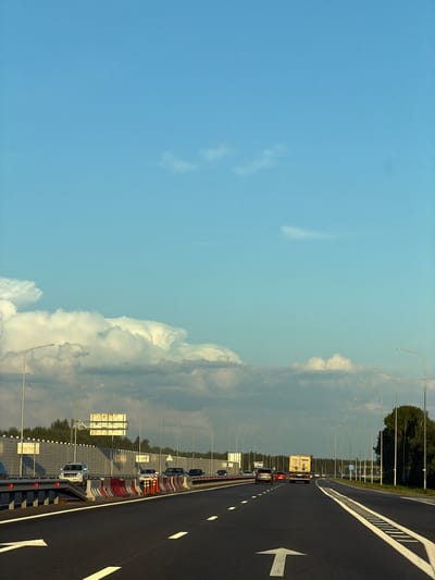 This is a vertical, eye-level, full shot of a highway in Zimnyaya Gorka, Russia, on a clear day with scattered clouds. The highway, which appears to be a multi-lane road, is paved with asphalt and marked with white lane lines. Several vehicles are visible, including cars, a truck, and a bus. The road is lined with streetlights and a sound barrier. Trees and vegetation are visible in the distance. The sky is a light blue, with white clouds.