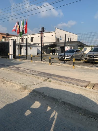The image depicts an exterior scene in Ikota, Nigeria, focusing on the entrance of a commercial or industrial compound under bright daylight.
In the foreground, a paved road with a concrete curb and an exposed metal drainage cover or ramp runs horizontally. The immediate road surface shows a pattern of concrete pavers, partially covered by dirt, and distinct shadows are cast from the left, indicating the sun's position.
The midground features a perimeter fence constructed of black metal mesh panels supported by black posts, secured by a series of yellow and black striped bollards across the entrance. Behind this barrier, a small, dark grey building, likely a security gatehouse or office, is visible, bearing a sign that reads "THREE ACE GROUP." Two Mercedes-Benz vehicles are parked within the compound, behind the fence: a silver C-Class sedan and a lighter-colored SUV.
The background is dominated by a large, white, multi-story building with a modern, angled roofline. To the left of this building, four flagpoles display the national flags of Canada, Belgium, Nigeria, and the United States, from left to right. A dark water tank on a tall, red-painted metal structure is situated behind the flagpoles. Overhead, power lines traverse the clear blue sky, which features sparse white clouds. No individuals are visible in the scene.