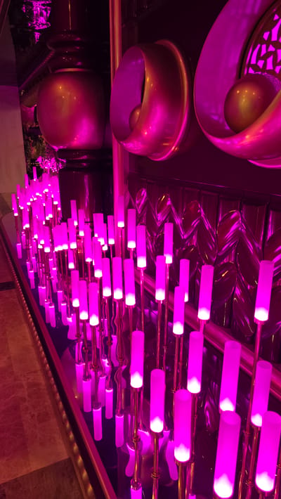 The image shows an indoor scene with abundant pink and purple lighting. In the foreground, a dense arrangement of tall, slender, illuminated rods in varying heights stand in rows. These rods are a vibrant pink and emit a bright white light from their upper sections. They appear to be a decorative lighting installation.

In the background, there are large, rounded, metallic objects with a gold or brass finish, illuminated by the pink lighting. Two of these objects are prominent, each appearing to be a hollow, funnel-shaped structure with a spherical object inside. Behind these, a textured wall or surface is visible, reflecting the pink light in a patterned way.

The floor in the foreground is covered in what appears to be marble or tiled flooring, with a warm, reddish-brown hue. The overall impression is one of a modern, possibly upscale interior, with a focus on dramatic lighting and decorative elements. The scene is clean and appears to be well-maintained. No people are visible in the image. The location is described as Kazafani, Northern Cyprus / Cyprus.