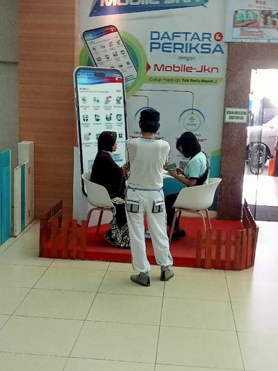 The image shows a scene inside a building in the Kecamatan Margorejo district of Surabaya, Indonesia (latitude: -6.76, longitude: 111.015). Three people are sitting at a table in front of a large advertisement. The ad is for a service called "Mobile-Jkn" and is decorated with a large digital phone display showing a list of icons. The phone is positioned to the left of the table. The person in the center is a man wearing a white shirt and white pants. He is facing the camera and looks to be talking to the woman sitting to his right. The woman has short, dark hair and is wearing a blue shirt. She is looking down at the table, presumably at a document. The woman seated to the left of the man is wearing a black headscarf and black dress. She is looking towards the phone. All three people appear to be interacting in some form of transaction. The scene is set on a raised platform, which has a wooden fence along the front. The floor is tiled, and the walls of the room appear to be a light color. There is an open doorway to the right of the scene which allows a glimpse into another room. The overall scene suggests an office or other business setting.
