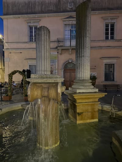 The image depicts a night scene in Anduze, France, focusing on a public fountain. The fountain is constructed with stone pillars and has water flowing from its upper sections into a basin. In the background, a building with a pale pink facade is visible. The building features architectural elements like windows with shutters and a balcony. To the left of the fountain, there is an arched structure adorned with foliage and flowers, under which tables and chairs are set up, suggesting an outdoor seating area for a cafe or restaurant. Several people are seated at the tables. A sign with text, likely advertising or information, is attached to the facade of the building. The overall environment appears to be a town square or public space.