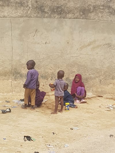 Four children are on the ground in Dutsinma A, Nigeria. The ground has a concrete section and a sandy, littered section. One child wears a maroon head covering, lying with another wearing a purple shirt and blue pants. Another child wears a purple shirt, lying on their stomach. The fourth child wears a purple shirt and tan pants and is standing.