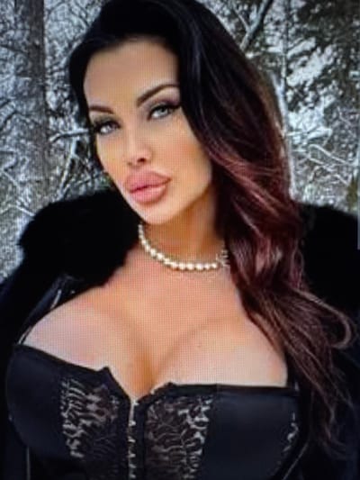 The image depicts a single female individual positioned in the city of Balykchi, Uzbekistan. The background is a blurry, desaturated outdoor setting, indicative of a wintry, possibly forested environment with visible tree trunks or branches partially obscured by snow or frost. The female subject has long, dark brown hair with visible reddish-brown highlights, styled to fall over her shoulders. Her face is fully made up, featuring prominent, full lips colored with light pink lipstick, precisely defined eyebrows, and heavily applied eye makeup accentuating her blue-green eyes, which maintain a direct gaze towards the viewer. She is wearing a single-strand pearl necklace. Her attire includes a black garment with a fur or faux-fur collar visible on her left shoulder and, beneath it, a low-cut black satin and lace corset or bustier with a central front closure, exposing her upper chest.