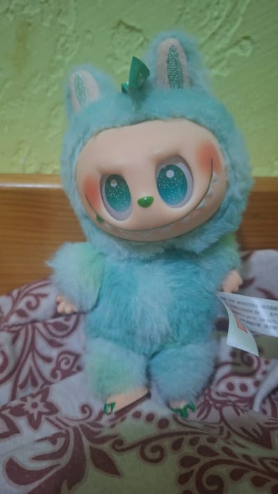 A plush toy is shown sitting on a patterned fabric. The toy is light blue with fuzzy fur and large, green eyes with white dots. It has small, pink cheeks and a wide smile showing tiny white teeth. A green bow is on top of its head. The background is a textured, light green wall. The fabric the toy is sitting on is patterned with brown swirls and floral designs. A white tag with text is attached to the toy's lower body. The scene appears to be indoors, possibly in a home environment. No specific location in the city of Voinova, Moldova can be determined from the image.