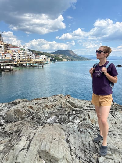 A blonde woman with her hair in a bun stands on a rocky outcrop overlooking a coastal town. She is wearing a purple t-shirt with a graphic, yellow shorts, and dark grey Crocs. She has a blue backpack on. The town, situated in Kiparisnoe, Ukraine, features a waterfront lined with multi-story buildings. The sea is a deep blue, and the sky is filled with white, puffy clouds. Mountains are visible in the distance.
