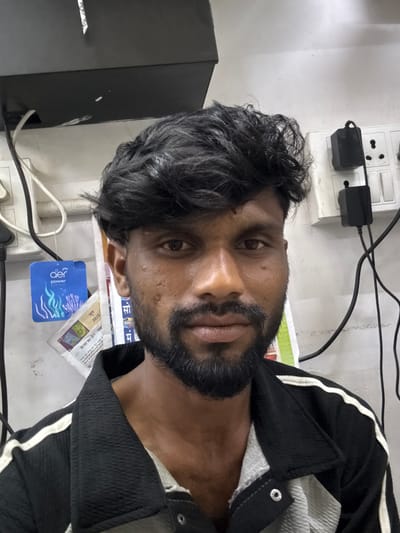 This image depicts an adult male in an indoor setting within the city of Palghar, India. The man has a dark complexion, dark, curly hair, and a full dark beard and mustache. He is looking directly into the camera. He is wearing a black jacket with a textured, crinkled fabric and white stripes along the shoulders, over a light-colored, possibly grey, textured shirt. Visible on his face are small skin blemishes or scars, particularly on his right cheek.
The background consists of a white wall that appears to be painted. On the left side of the man, a blue "aer power pocket" air freshener is hanging, displaying "power pocket" and "jun 2025" (June 2025). Behind and below the air freshener, several papers or documents, possibly newspaper clippings with Hindi text, are affixed to the wall. Various electrical cables, black and white, are visible, some running vertically and horizontally.
On the right side of the man, a white electrical outlet board with multiple sockets and switches is mounted on the wall. Two black power adapters are plugged into the sockets, with numerous black electrical cables extending from them and across the wall. Above the man's head, a black shelf or surface is visible, with a small white sticker or piece of paper attached to its underside and some white specks on its surface. The environment suggests a functional space, possibly a utility area or a simple room, characterized by visible electrical infrastructure and functional elements.