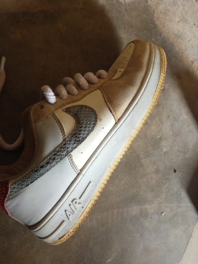 A close-up, top-down shot shows a single, tan and white Nike Air Force 1 sneaker with a snake-print swoosh. The sneaker is resting on a textured, grey concrete surface. The laces are a light tan color and are loosely tied. The sneaker appears to be slightly worn, with some discoloration on the sole. The background is out of focus, with the dominant feature being the sneaker. There are no people visible in the image. The context provided about Gindin Dutse, Nigeria is not visually supported by the image content.