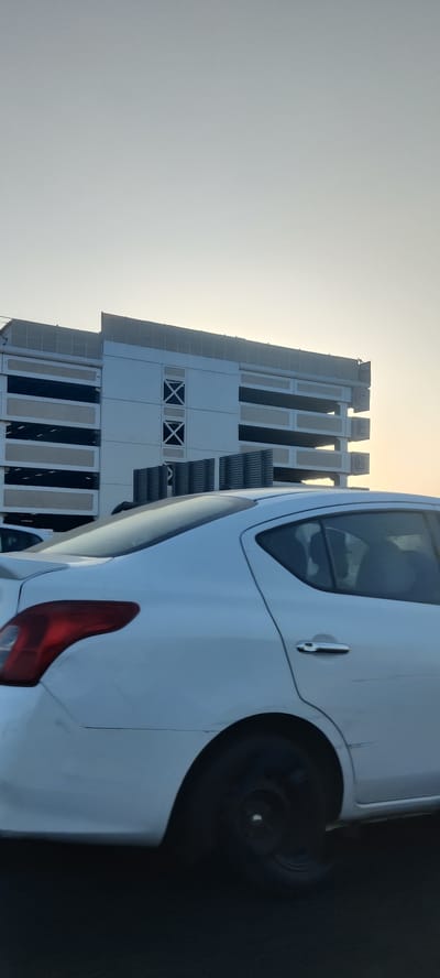 A white sedan is positioned in the foreground, likely captured from another vehicle. Its rear tire is visible, appearing dark and blurry due to motion. The body of the car shows some minor scratches and imperfections. Behind the car is a multi-story parking garage. A sign marked "CEB" is fixed on the side. The sky appears hazy, with a light color gradient suggesting either sunrise or sunset. The location is likely Jeddah, Saudi Arabia, considering the context.