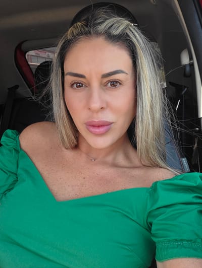 The image is an angled, close-up portrait of a woman with blonde hair. She is looking directly at the camera with a neutral expression. She is wearing a green off-the-shoulder top with ruffled sleeves. A thin silver necklace with a small diamond pendant is visible on her neck. The background suggests she is inside a car, with a glimpse of the car's interior, including part of a car mirror. The lighting indicates it is daytime. There is text visible on a part of the car's interior, which reads "AUDIA" in reverse.
