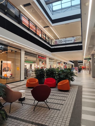 The image depicts the interior of a modern shopping mall in Zielona Góra, Poland. The scene shows a wide, well-lit corridor with shops on either side and a multi-level structure above. In the foreground, there are several modern, brightly colored chairs – orange, red, and maroon – arranged in a seating area on a patterned tile floor. A small, white rectangular object, possibly a planter or a speaker, is situated between two chairs.
The shops visible include "Fielmann" (an optician) with large glass windows displaying eyewear and advertising, and another shop with signage indicating a sale with "30%" discounts. Further down the corridor, more shops are visible, as well as a directional sign pointing towards "Parking P" and an exit. The architecture features sleek lines, ample artificial lighting, and a skylight system at the top of the mall. Several large potted plants are strategically placed throughout the seating area, adding a touch of greenery. In the background, a few shoppers are casually walking through the mall. The overall impression is one of a clean, contemporary retail environment.