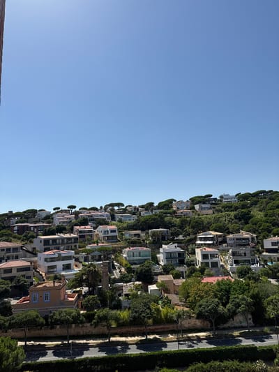 This is a vertically oriented, wide-angle, eye-level shot of a dense collection of modern villas nestled into a steep hillside. The buildings are a mix of white, beige, and grey, with large windows and flat roofs, many adorned with solar panels. Lush green trees and manicured hedges surround the homes, with a paved road visible to the right, lined with streetlights and casting long shadows. The scene appears to be in a Mediterranean climate, suggested by the vegetation and architectural style. The sky is a clear, bright blue, indicating a sunny day, likely late morning or early afternoon due to the angle of the shadows. No people are visible, and no text is present. The overall impression is one of affluence and a harmonious integration of modern architecture with a natural, hilly landscape. The location cues of Sant Feliu De Guíxols, Spain, further confirm the Mediterranean setting.