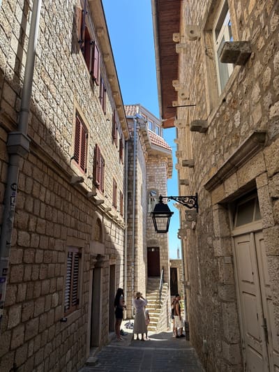 The image depicts a narrow street in the old town of Ulcinj, Montenegro. The street is paved with dark cobblestones and is flanked by stone buildings on both sides. The architecture is characterized by rough-hewn stone walls, red-tiled roofs, and wooden shutters.
On the left side of the street, a stone building with visible brickwork features several windows with closed red shutters. A grey downspout runs vertically along the wall, adorned with several stickers.
On the right side, another stone building presents a lighter stone facade with a large white door. Above the door, there is a stone ledge and a window. A decorative black metal lantern is attached to the wall by an ornate bracket.
In the middle of the street, a group of people are visible. Two women and a child are standing on the left side, near a doorway with a stone arch. One woman, wearing a straw hat and a long white dress, is looking towards the right. The other woman, in a dark top and skirt, is facing away from the camera. A child in a pink dress stands beside them.
Further down the street, on the right side, a man and a woman are standing. The man is wearing a dark t-shirt, shorts, and a cap, while the woman, also wearing a straw hat and a light-colored dress, is facing the man.
The street recedes into the distance, leading to a set of stairs and another building with more windows and a distinctive tiled roof. The sky above is clear and blue. The overall scene conveys a sense of historical architecture and a leisurely atmosphere in a Mediterranean town.