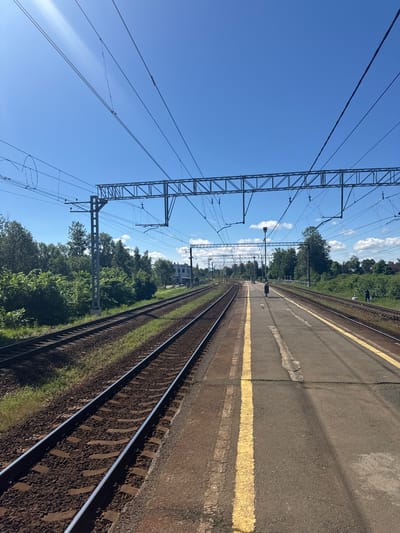 The image depicts a railway station platform in Kobrovo, Russia, on a clear, sunny day. The foreground shows two sets of train tracks running parallel. The platform itself is paved and marked with a bright yellow line indicating the edge. In the mid-ground, several people can be seen walking along the platform. Overhead, a complex network of electrical wires and support structures for the railway is visible against the blue sky. Beyond the tracks and platform, lush green trees and vegetation line the landscape, with a few buildings and more trees visible in the distance. The scene conveys a sense of an active, albeit quiet, suburban or rural railway station.