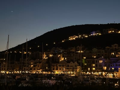 The image depicts a coastal town at dusk or night, with buildings nestled on a hillside overlooking a harbor filled with sailboats. The sky is a deep blue, transitioning to a lighter shade near the horizon. A single bright star or planet is visible in the upper left quadrant of the sky. The buildings on the hill are illuminated by warm yellow lights, creating a cozy and inviting atmosphere. The harbor is dotted with numerous masts of sailboats, some of which are also lit. The overall scene suggests a serene and picturesque evening in a Mediterranean setting. This specific location is identified as Gošići, Montenegro.