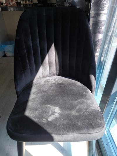 An indoor shot shows a black velvet chair positioned in front of a window with blue light filtering through. The chair's seat has several light-colored smudges, possibly dust or lint, concentrated in the center. A shadow from the window bisects the chair, with a portion of the seat and backrest illuminated by sunlight. To the left of the chair, a blue plastic trash bag is visible inside a patterned waste bin. The floor is a light-colored wood grain. The background includes a countertop with various containers and a patterned wall. The scene appears to be located in Pendik, Turkey.