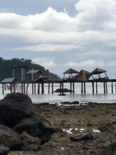 This image depicts a dilapidated wooden pier extending into the ocean in Layeun, Indonesia. The pier is in a state of disrepair, with several small, open-sided shelters or gazebos on it. Some of these structures have collapsed or are heavily damaged, with their roofs tilted or missing. The wooden pilings supporting the pier are visible, some of which appear worn or partially submerged. The water is calm, reflecting the overcast sky, which is filled with a mixture of white and gray clouds. The foreground shows a rocky and pebbly shore, with several large, dark rocks scattered along the beach. In the background, a lush, green hillside with dense vegetation rises behind the pier. The overall scene conveys a sense of abandonment and decay, with nature reclaiming the man-made structure. There are no people visible in the image.