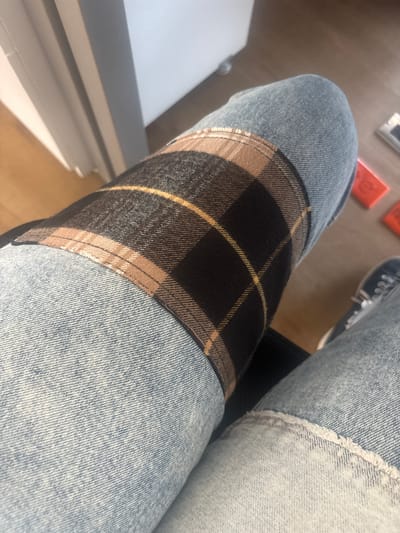 This close-up image, set in Buenavista, Mexico, features a person's legs clad in light-wash denim jeans, seen from a high-angle perspective. The individual appears to be seated or resting their legs in an indoor environment. A notable detail on the jeans is a large, rectangular plaid fabric panel sewn onto the knee of the left leg (from the viewer's perspective). This plaid pattern is composed of dark, possibly black or deep brown, interwoven with intersecting stripes of light brown/beige and thin golden-yellow lines. The stitching around this patch is clearly visible.
In the background, a warm-toned wooden floor is visible, sparsely adorned with a few small, light-colored rectangular objects and some red items. The presence of strong highlights and shadows indicates bright, natural daylight, suggesting it is a sunny day. On the right side of the frame, the front portion of a shoe, likely a sneaker with white laces, is partially visible. To the left, a segment of a white or light-colored furniture piece can be seen. No specific activity is occurring, and there is no visible text or discernible emotions.